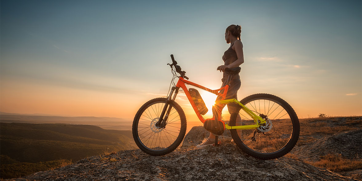woman on cliff edge with electric bike looking out over landscape at sunset